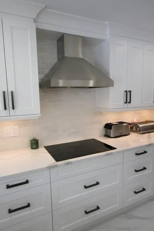 Modern kitchen with a stainless steel hood above a black induction cooktop, surrounded by white cabinetry and quartz countertops, featuring a toaster and bread box on the right.