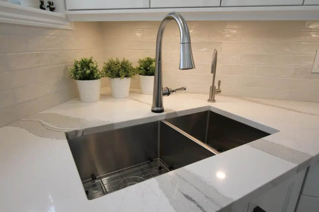 Modern kitchen sink with a sleek stainless steel faucet, white quartz countertop, and three small potted plants in the background against a beige tiled backsplash.