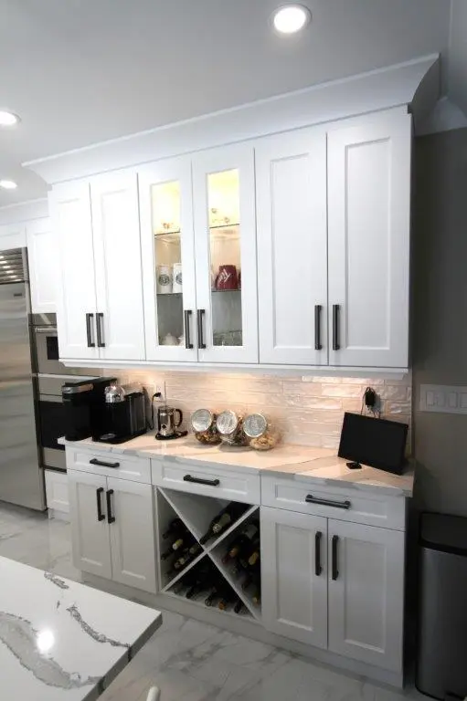 A modern kitchen cabinet setup with white cabinets and black handles. The upper cabinets feature glass doors and are lit from the inside, displaying decorative items. The lower cabinets include a marble countertop, a coffee machine, and organized storage for wine bottles. A digital device, possibly a tablet, is propped up on the counter.