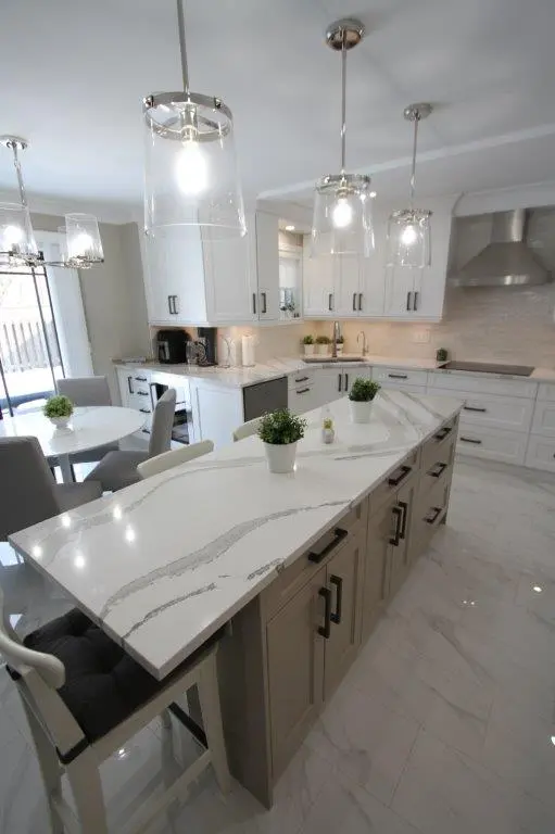 Modern kitchen with a large white quartz island, pendant lights, white cabinets, stainless steel appliances, and a small dining table with gray chairs.