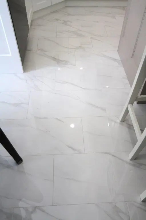A kitchen floor with shiny white marble tiles featuring subtle gray veining, reflecting overhead lights.