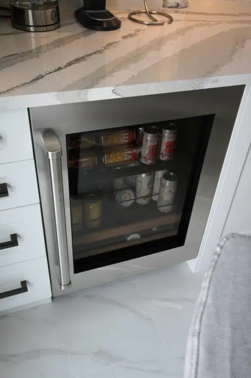 A small beverage fridge built into a kitchen cabinet, containing several cans of drinks visible through the glass door.