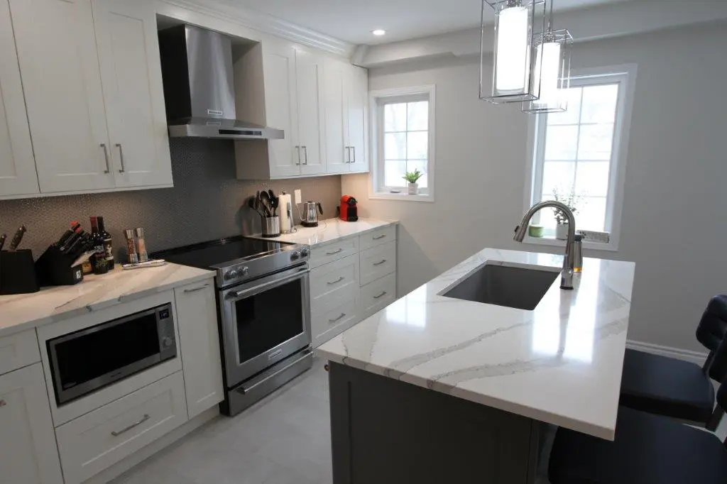 Modern kitchen with white cabinetry, stainless steel appliances, a quartz countertop, an island with a sink, and pendant lighting, featuring two windows providing natural light.