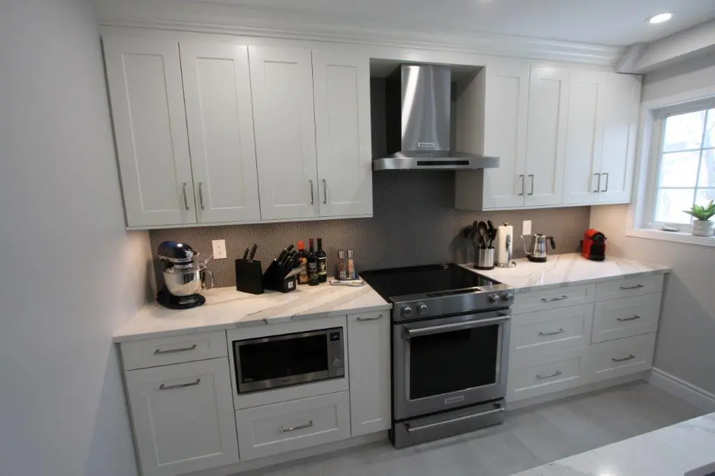 Modern kitchen with white cabinets, a stainless steel oven and range hood, quartz countertops, and a window with a small plant on the sill. Various kitchen appliances and utensils are placed on the counter, including a mixer, knife block, and coffee machine.