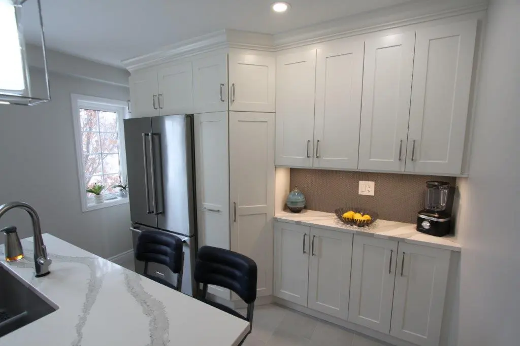 Modern kitchen with white cabinetry, a stainless steel refrigerator, a countertop with a vase and a blender, and two padded bar stools by a breakfast bar.