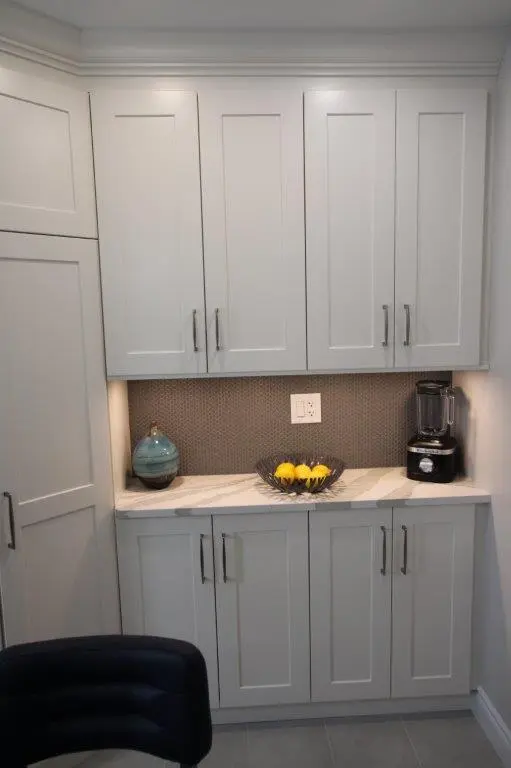 A kitchen corner featuring white cabinets with silver handles, a quartz countertop, and a textured backsplash. It includes a small decorative bowl of lemons in the center, a blue vase on the left, and a blender on the right, with under-cabinet lighting illuminating the space.