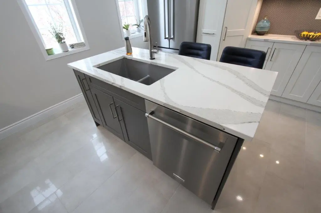 Modern kitchen island with a white marble countertop featuring gray veining, an under-mount sink, and a sleek stainless steel dishwasher beneath. The surrounding area includes a light grey floor and white cabinets with decorative items on display.