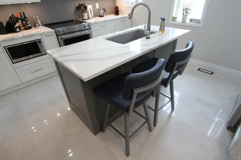 A modern kitchen with a central island featuring a white countertop with gray veining, a built-in sink, and a tall faucet. Two dark-colored chairs are positioned at the island, facing the sink. The surrounding kitchen cabinets are white, and there is a stainless steel stove and microwave installed in the cabinetry. A window with small potted plants is visible in the background.