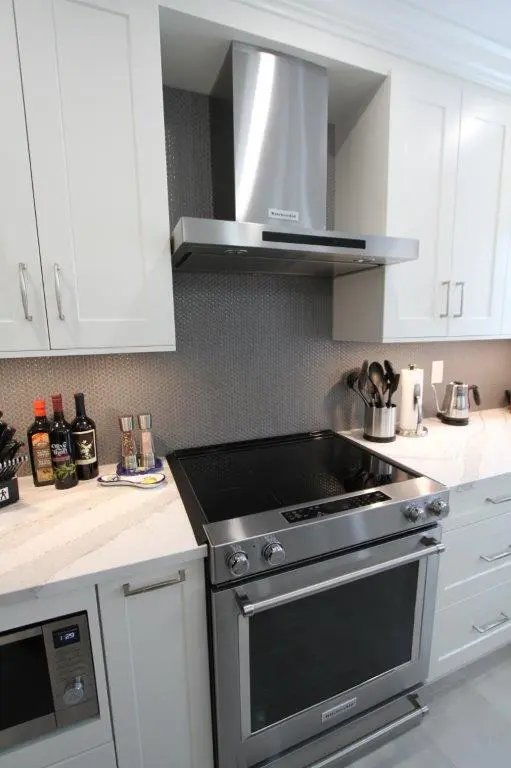 Modern kitchen with a stainless steel stove and range hood, white cabinets, a speckled gray backsplash, and marble countertops featuring various kitchen utensils and bottles on the counter.