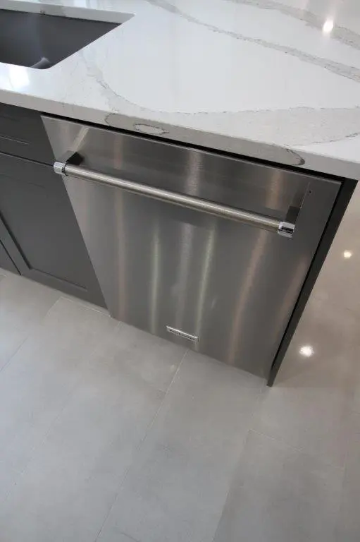 A modern stainless steel dishwasher installed under a white marble countertop in a kitchen with gray cabinets and tile flooring.