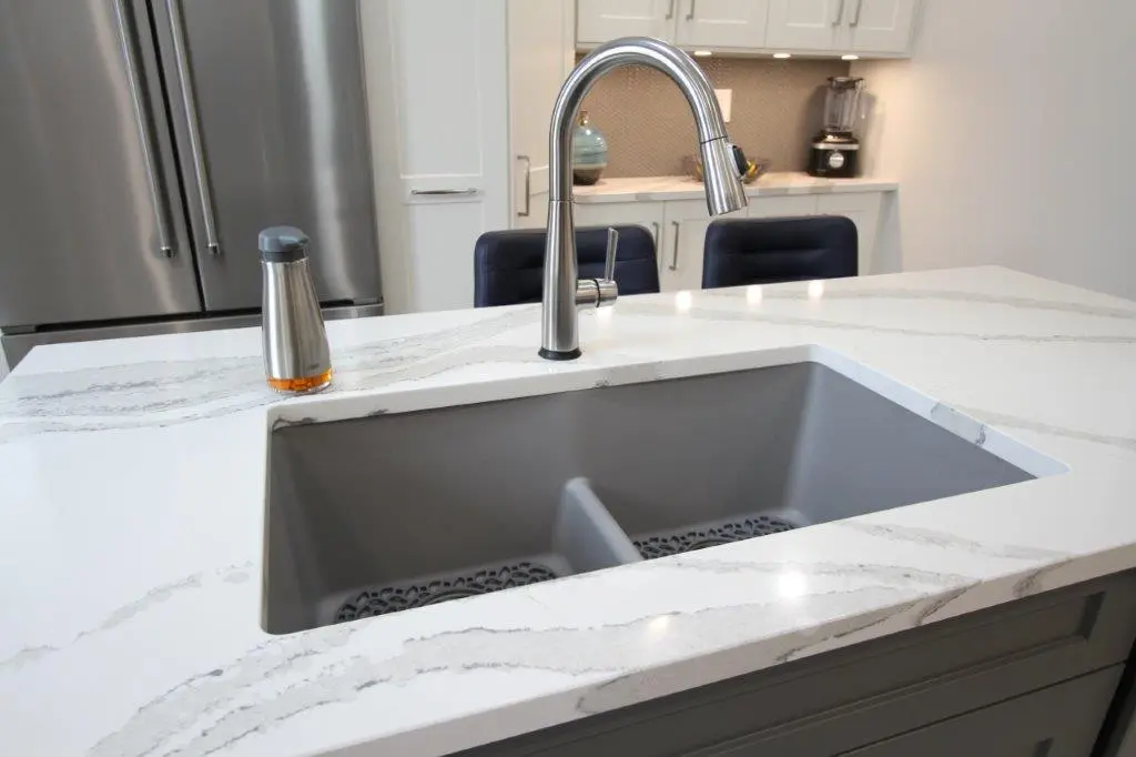 A modern kitchen sink with a stainless steel faucet, set within a white marble countertop featuring gray veining. A soap dispenser is adjacent to the sink, and stainless steel kitchen appliances are visible in the background.