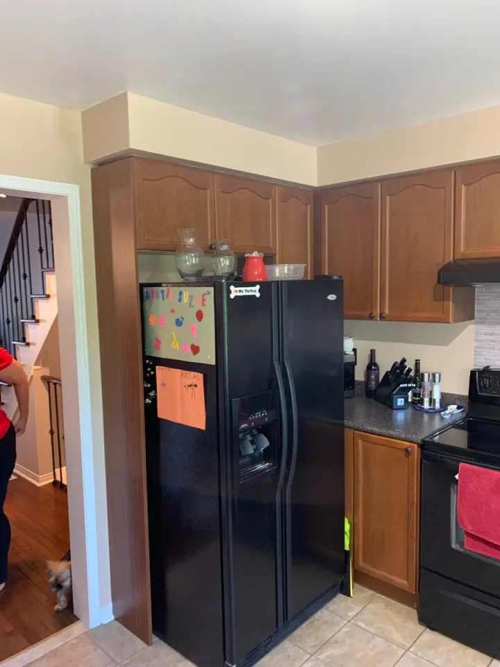 A before renovation kitchen with wooden cabinets, a black refrigerator with children's drawings and magnets, a black stove, and various kitchen items on the countertop.