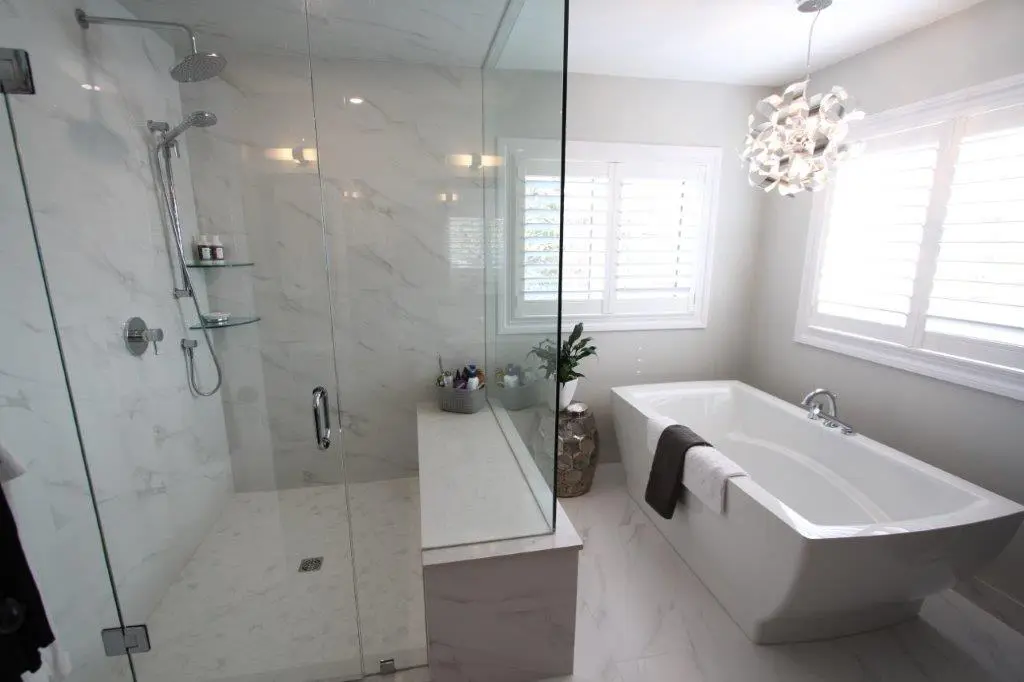 Modern bathroom with a large glass-enclosed shower and white tiled walls, featuring a freestanding bathtub next to a window with white blinds, and a unique pendant light overhead.