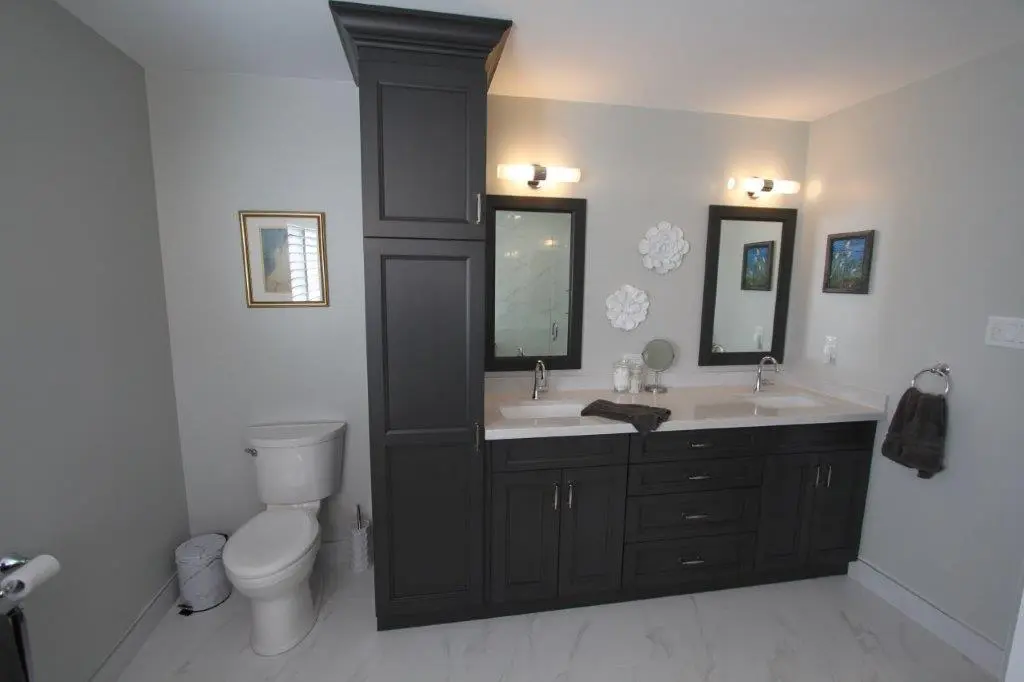 A modern bathroom with a dual sink vanity featuring dark cabinetry, two wall-mounted mirrors, and two light fixtures. The toilet is positioned to the left, with light-colored walls and tiled flooring. Decorative wall art and a towel ring are on the walls.