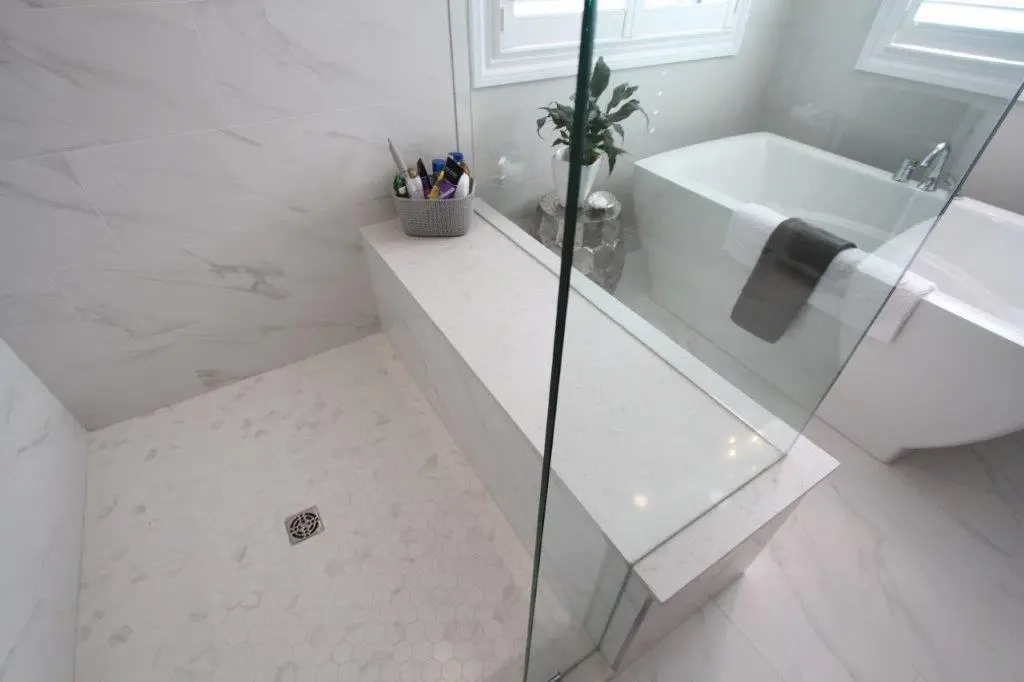 A modern bathroom featuring a corner shower with a marble bench, glass enclosure, hexagonal floor tiles, and a freestanding bathtub with a towel draped over the side.