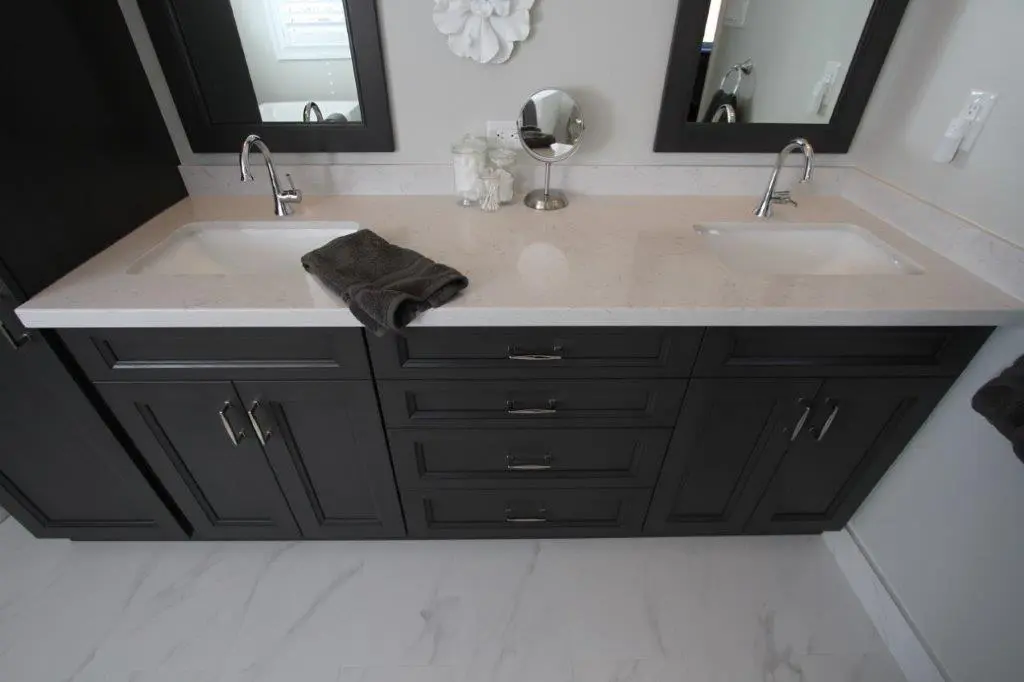 A modern bathroom vanity with a double sink and dark cabinetry, featuring a light countertop. A small mirror and a jar are placed between the sinks, and a folded towel is draped over one of them. The floor is covered with light-colored tiles.