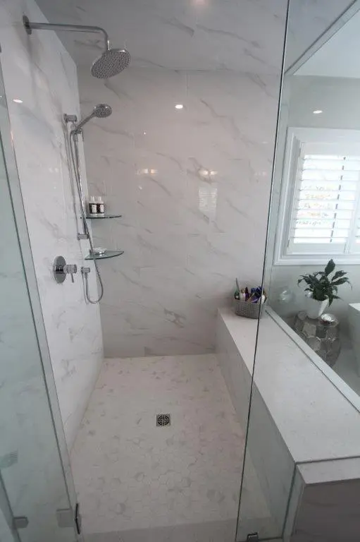 A modern bathroom with marble-patterned tiles and a glass-enclosed shower featuring a rain showerhead. The floor has a hexagonal tile design, and there is a built-in bench with toiletries. A window with shutters is in the background beside a small plant.