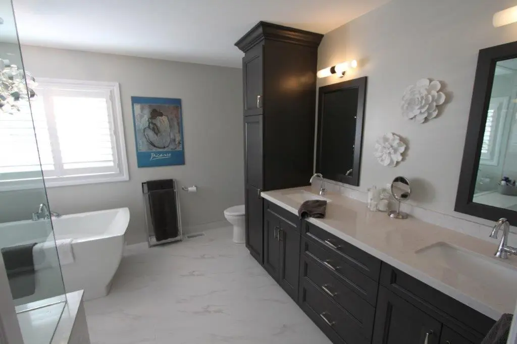 Modern bathroom with dark cabinetry, a double vanity with sinks, wall-mounted light fixtures, a free-standing bathtub near a window, and a piece of framed art on the wall.