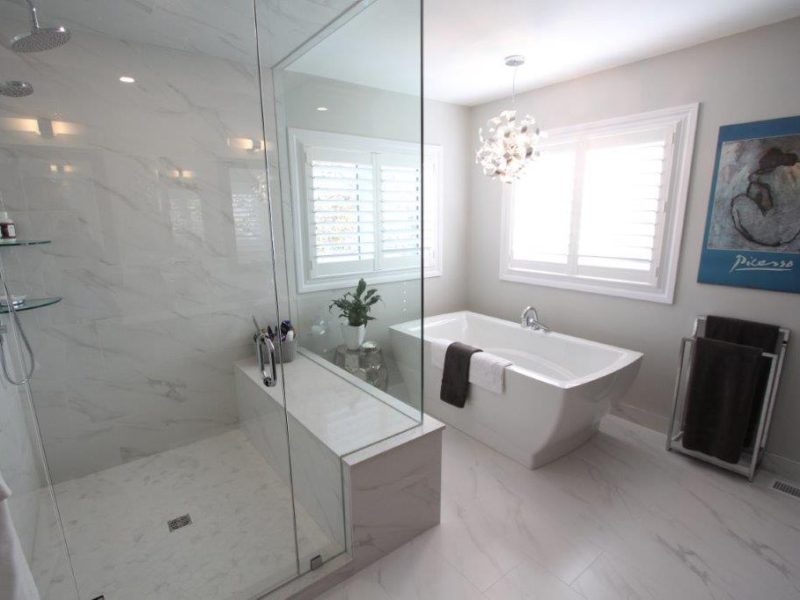Modern bathroom featuring a glass-enclosed shower with marble-like walls, a freestanding bathtub next to a window with white shutters, a decorative pendant light, and a painting on the wall.