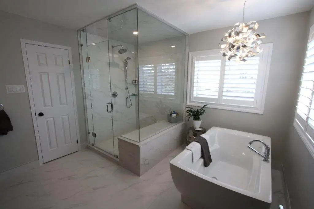 A modern bathroom with a large glass-enclosed shower on the left, featuring a marble floor and walls. To the right is a white freestanding bathtub with a silver faucet, next to a window with white shutters. A contemporary light fixture hangs from the ceiling, and a potted plant is placed on a small stool beside the tub.