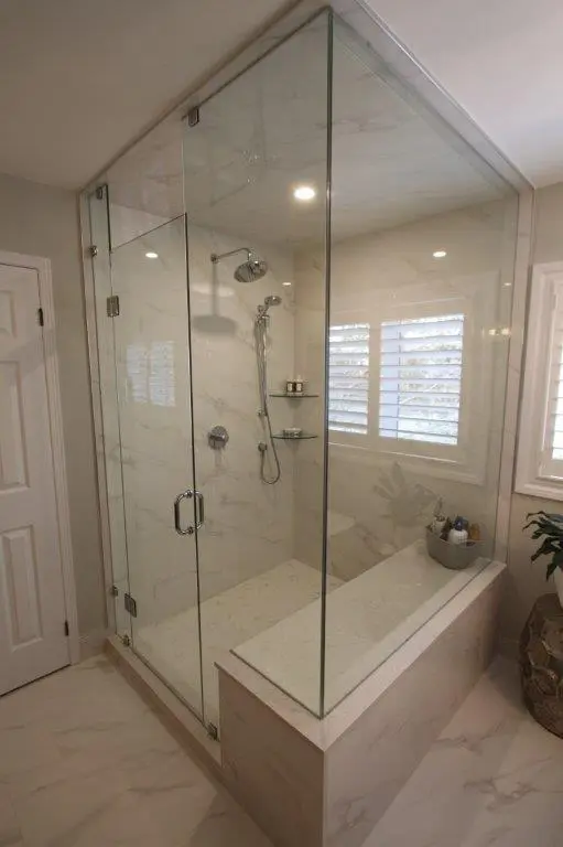 A modern bathroom featuring a glass-enclosed shower with marble walls and a built-in bench. The showerhead and fixtures are mounted inside, and there is a window with shutters on the adjacent wall. The floor and bench have matching marble finishes.