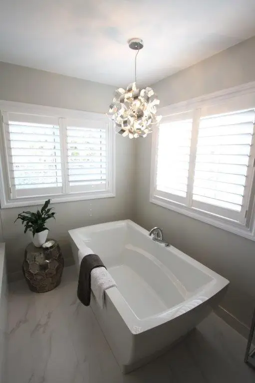Modern bathroom with a rectangular white bathtub beneath a contemporary white pendant light, surrounded by windows with shutters, and a small decorative table with a potted plant on top.