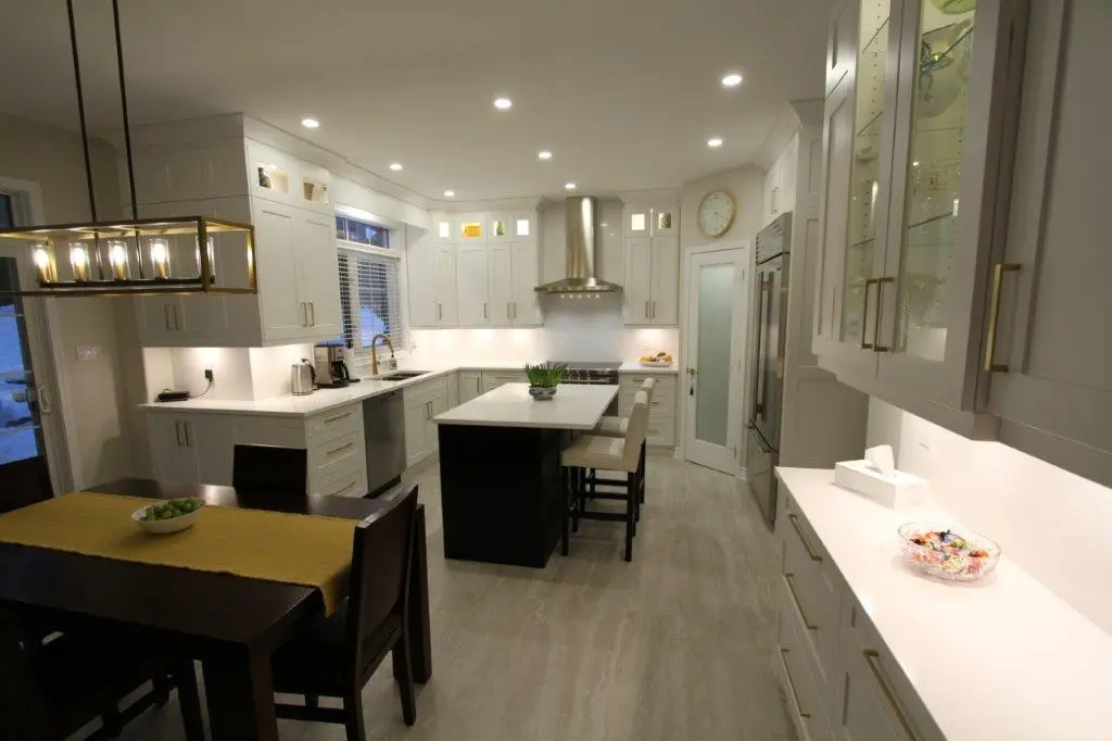 Modern kitchen with white cabinets, a central island with bar stools, stainless steel appliances, and recessed lighting, featuring a dining table with a yellow runner and decorative bowl.