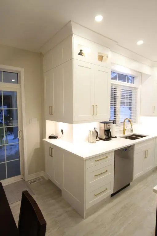 Modern kitchen featuring white cabinetry, a stainless steel dishwasher, a coffee maker, and a silver faucet under a large window with blinds. The room is well-lit with recessed lighting and has a sliding glass door leading outside.