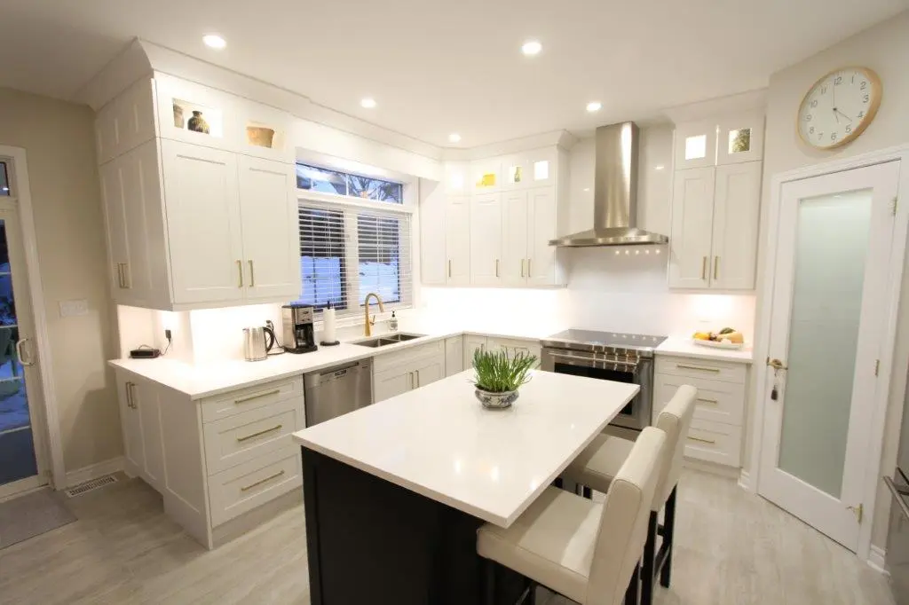 Modern kitchen with white cabinetry, a central island featuring two bar stools, a stainless steel stove, and a range hood, illuminated by recessed lighting.