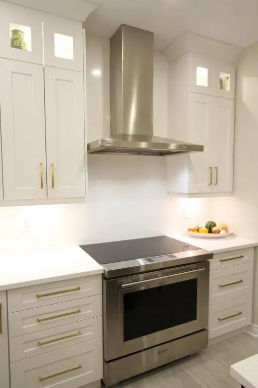 Modern kitchen with white cabinets, a stainless steel stove, and a range hood, featuring gold hardware and a display of vegetables on the countertop.