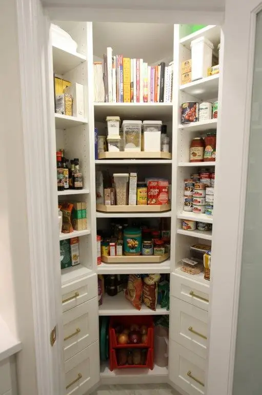 A neatly organized pantry with shelves filled with various food items, including spice jars, canned goods, cereal boxes, and jars. Cookbooks are stacked on the top shelf, while storage bins and containers hold onions and potatoes at the bottom. The pantry has white shelves and cabinets with gold handles.