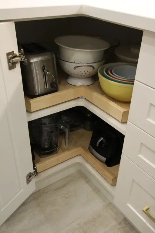 A corner kitchen cabinet with open doors revealing two shelves. The top shelf holds a toaster, a white colander, and a stack of colorful bowls. The lower shelf contains a blender and a black appliance, possibly a coffee maker or air fryer.