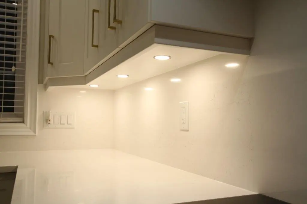 A modern kitchen interior featuring white cabinetry with gold handles, under-cabinet lighting, and a white quartz countertop and backsplash. There are electrical outlets and a window with blinds on the left side.
