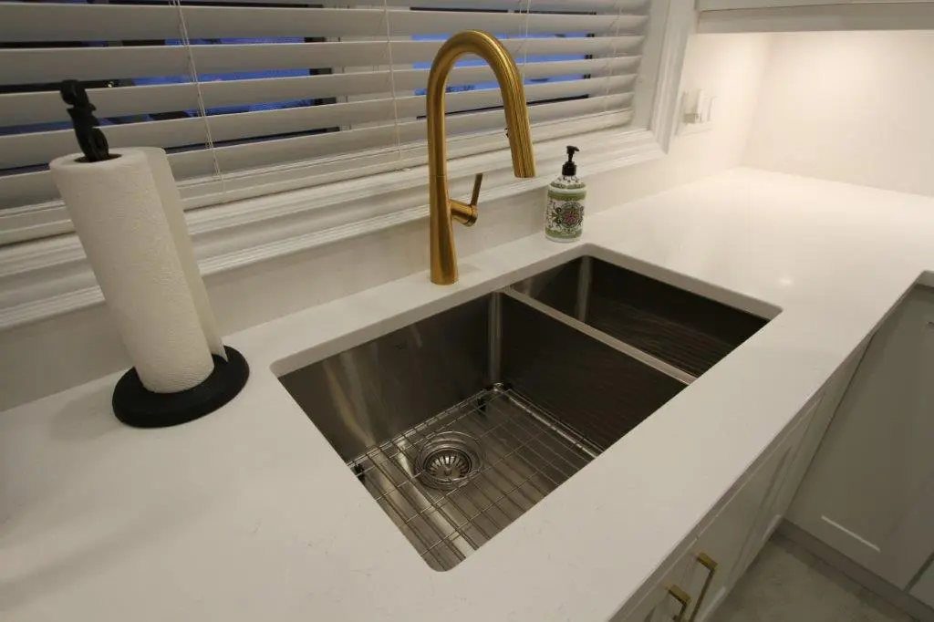 A modern kitchen sink with a gold faucet, adjacent to a roll of paper towels on a black holder and a bottle of soap on a white countertop, with blinds covering the window in the background.