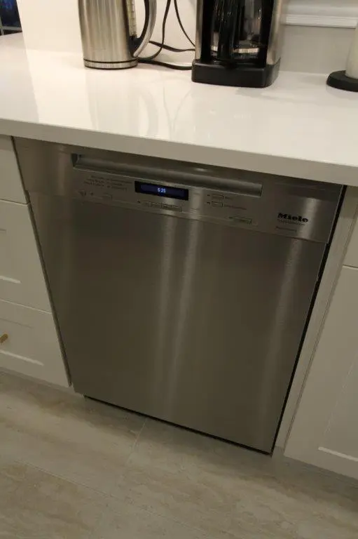 A stainless steel Miele dishwasher fitted under a kitchen countertop next to white cabinetry, with an electronic display panel on top.