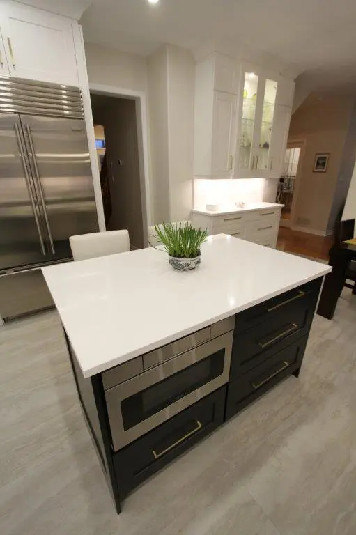 Modern kitchen with a white countertop island featuring built-in drawers and a microwave, a stainless steel refrigerator, and white cabinetry in the background.
