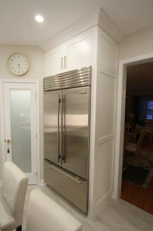 A modern kitchen with a stainless steel double-door refrigerator fitted into white cabinetry. The kitchen features a frosted glass pantry door and a wall clock. A portion of a dining room is visible through an open doorway.