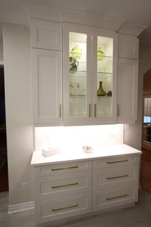 A white kitchen cabinet with glass upper doors displaying decorative glassware and green vases, and a countertop with drawers underneath, illuminated by under-cabinet lighting.