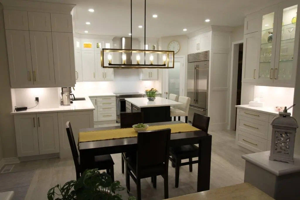 A modern kitchen with white cabinetry, stainless steel appliances, and a central island illuminated by a large rectangular chandelier. A wooden dining table with dark chairs is in the foreground, and the room features recessed lighting and a clock on the wall.