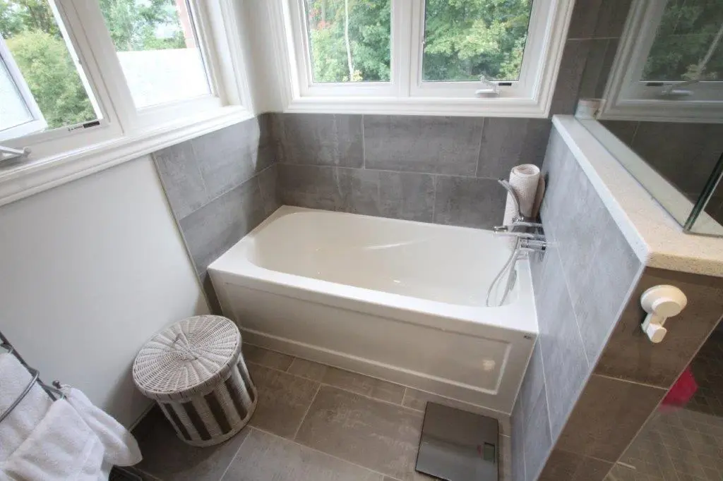 A bathroom featuring a white bathtub fitted into a corner with gray tiled walls and floors. The room is well-lit from windows above the tub, and there's a wicker basket and a digital scale on the floor nearby.