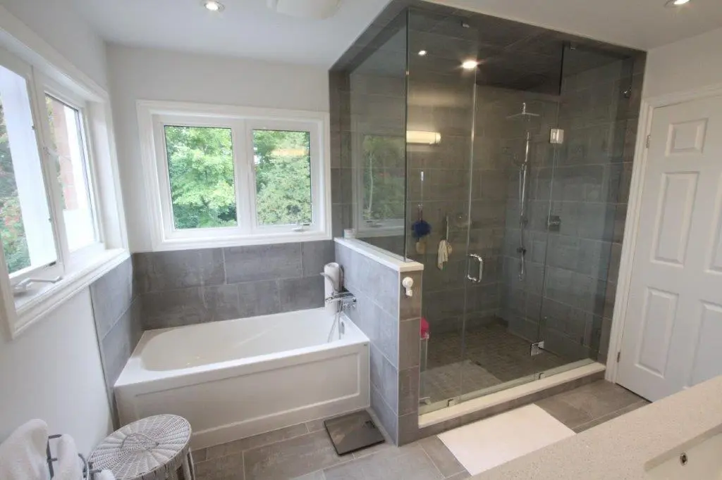 Modern bathroom featuring a glass-enclosed shower with a gray tiled interior and a white bathtub situated next to two large windows, complemented by a gray and white color scheme.