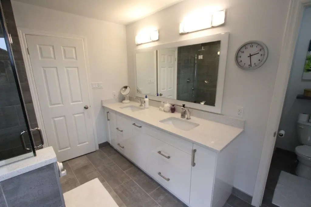 A modern bathroom featuring a double vanity with white countertops and drawers, a large mirror, overhead lighting, a wall clock, and a glimpse of a glass shower and toilet area.