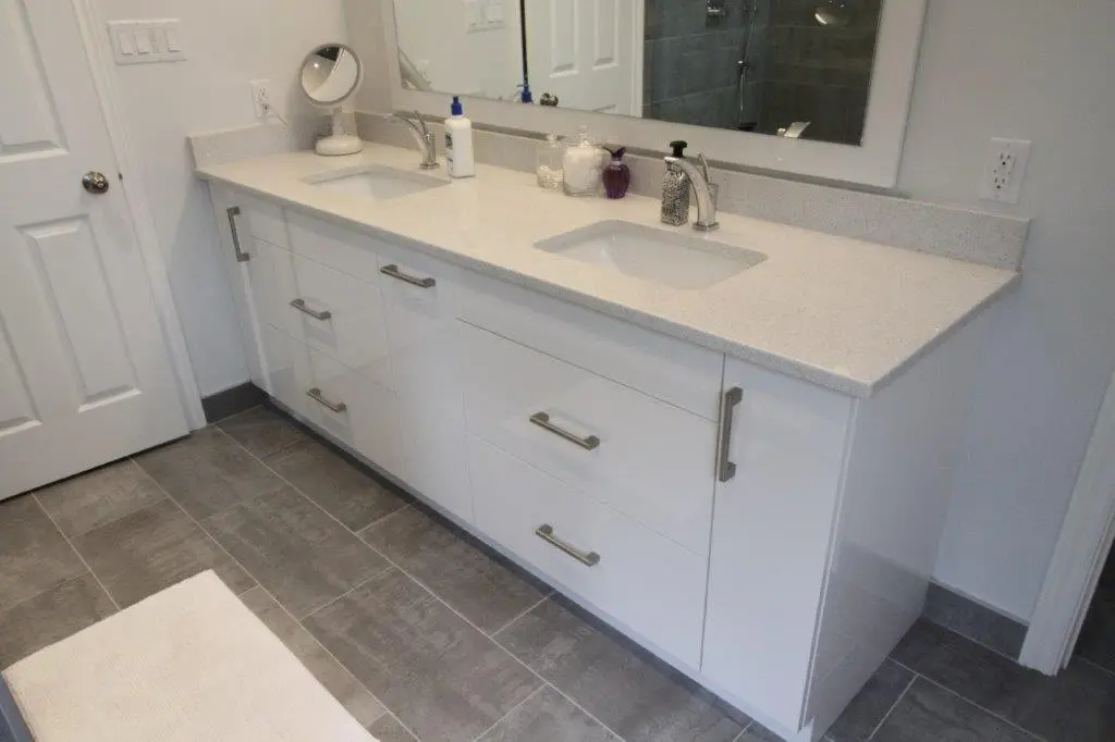 A modern bathroom with a white dual-sink vanity featuring metallic handles, a large mirror above, gray tile flooring, and various toiletries on the countertop including a soap dispenser and decorative containers.