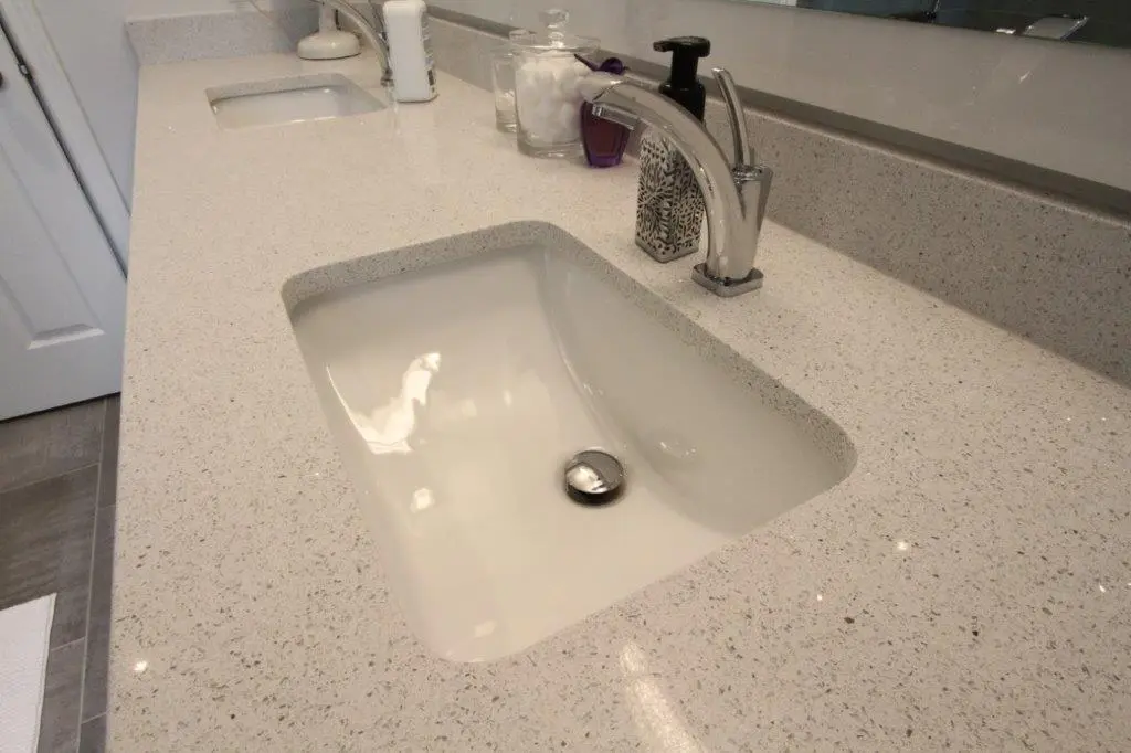 A bathroom countertop with an integrated rectangular sink and a chrome faucet. The countertop is light-colored with small specks and has toiletries such as soap dispensers and a toothbrush holder arranged on it.