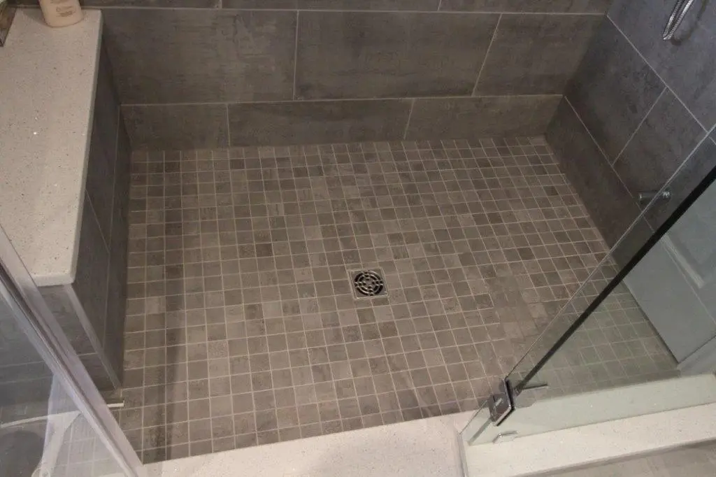 A modern walk-in shower with gray square mosaic floor tiles, a central drain, and glass doors.