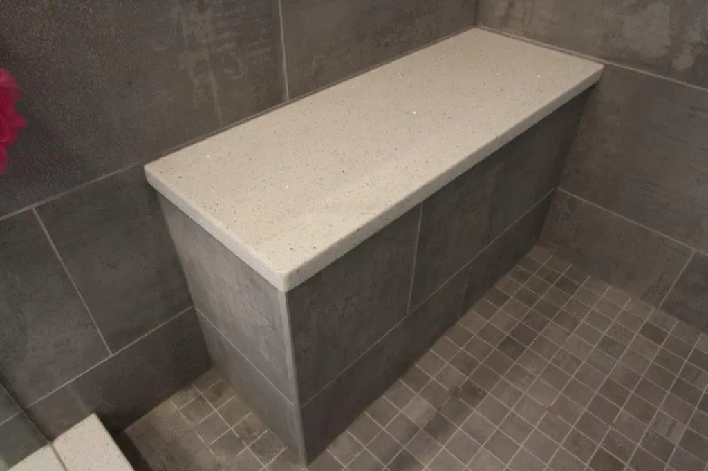 A bathroom with a corner bench made of gray tiles and a smooth beige top, set against tiled walls and floor.