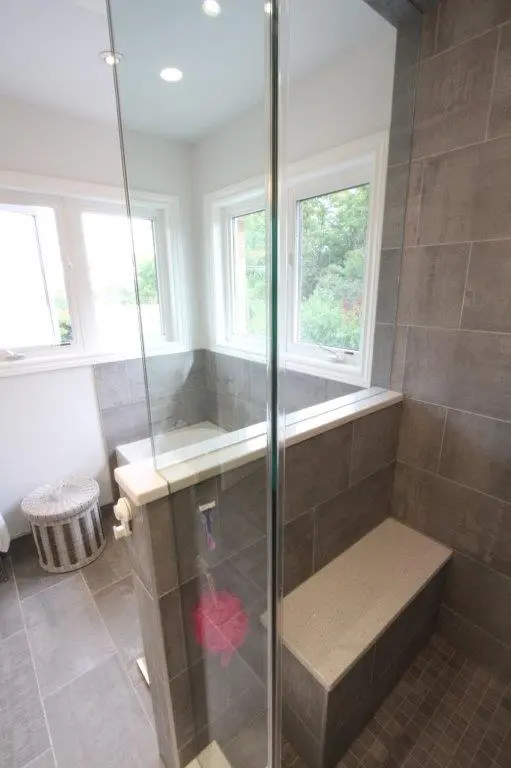 A modern shower with gray tiled walls and a built-in bench, featuring a glass door and two windows providing natural light.