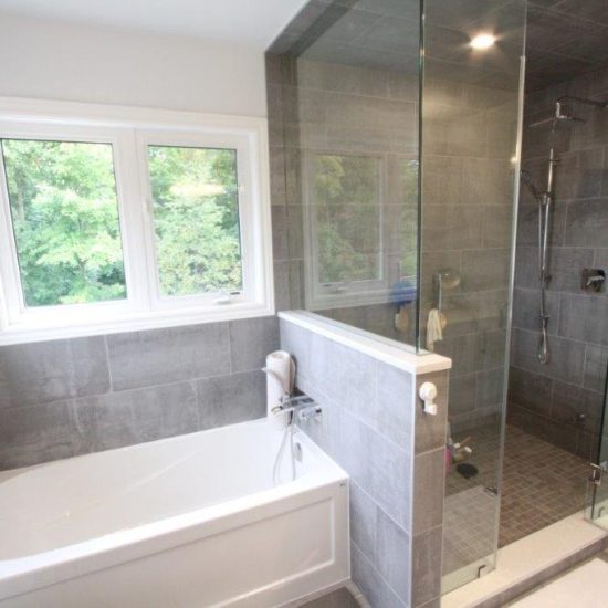 Modern bathroom with a white bathtub next to a large glass-enclosed shower area, set against gray tiles and a window overlooking greenery.