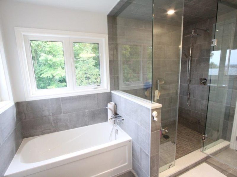 Modern bathroom with a white bathtub next to a large glass-enclosed shower area, set against gray tiles and a window overlooking greenery.