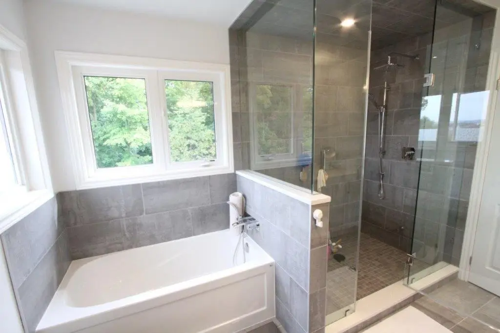 Modern bathroom with a white bathtub next to a large glass-enclosed shower area, set against gray tiles and a window overlooking greenery.